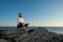 Side View Of Young Female In Warm Activewear And Knitted Hat Meditating With Mudra Hand Gesture While Sitting On Rock Against Blue Sky