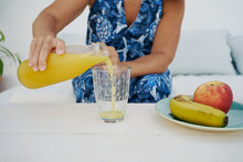 Crop Unrecognizable Female In Summer Dress Pouring Fresh Orange Juice From Bottle Into Glass Placed On Table With Fruits At Home