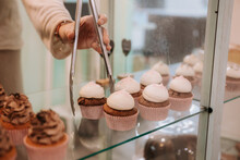 Crop Anonymous Female Seller With Tongs Taking Delicious Cupcake With White Cream From Glass Display While Working In Confectionery Shop