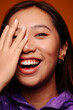 © Victor Torres/ADDICTIVE STOCK - Close-up portrait of cheerful Asian young woman. She wears a purple jacket and is looking at camera smiling with hand on face against a yellow background