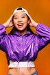 © Victor Torres/ADDICTIVE STOCK - Portrait of happy Asian young woman. She wears a purple jacket and a grey cap and is looking at camera smiling against a yellow background