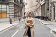 © The Trio Studio/ADDICTIVE STOCK - Thoughtful young African American female in trendy coat listening to music in true wireless earbuds while walking on London street with laptop in hand