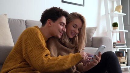 Wall Mural - Young happy couple having fun holding smartphone using mobile phone at home, laughing, playing game, doing online shopping in ecommerce app together, checking social media applications together.