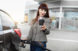 © sofiko14 - Transportation, technology, energy concept. Blurred portrait of young pretty businesslady, refueling her car at gas station, and demonstrating to camera paper cup of coffee to go. Focus on the cup