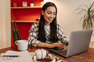 Brown-eyed woman in plaid jacket with smile working in laptop and posing at table with cup of coffee