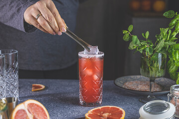 Wall Mural - Real woman preparing pink Grapefruit Mezcal Paloma Cocktail in highball glass with water drops on table in real home. New life concept of quarantine