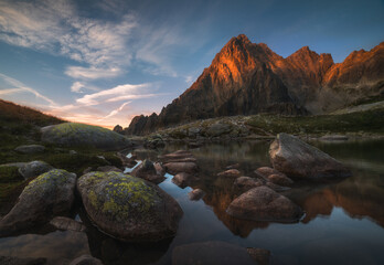 Wall Mural - Sunlit Rocky Mountain Peak and Mountain Lake with Rocks in Foreground at Sunrise. Mala Studena Valley, High Tatra, Slovakia.