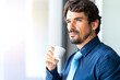 © Joao - Business man smiling and looking through the window holding a cup of coffee. Success man portrait with serious face