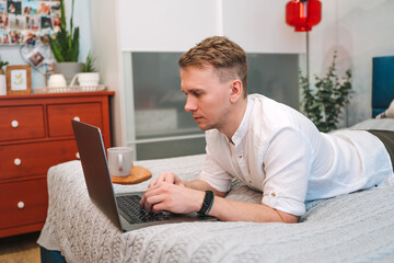 Canvas Print - a handsome man in a shirt uses a laptop while lying on the bed. the concept of the home office and o