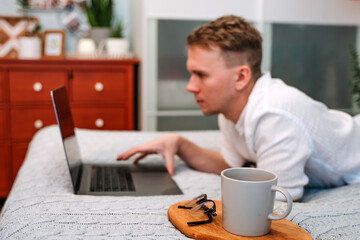 Canvas Print - a handsome man in a shirt uses a laptop while lying on the bed. the concept of the home office and o