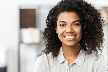 Headshot of a young elegant African American ethnic female with Afro curly hairstyle, beautiful smile and looking at the camera while standing against blurred home or office interior background