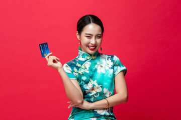 Wall Mural - Smiling happy Asian woman wearing oriental qipao dress showing credit card in hand on red studio background for Chinese new year cashless spending concepts