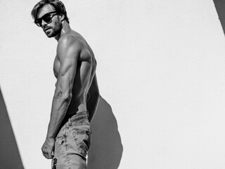 Portrait of sporty handsome strong man. Healthy athletic fitness model posing near white wall in jeans. Confident sexy fashion male with naked nude torso. Lambersexual outdoors in sunglasses