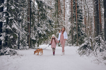 Happy family young mother and little cute girl in pink warm outwear walking having fun with red shiba inu dog in snowy white cold winter forest outdoors. Family sport vacation activities.