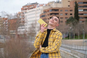 Wall Mural - Handsome young man wearing make up, laughing recording an audio voice message on his smartphone. Non binary androgynous guy.