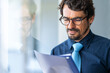 © Joao - Business man wearing glasses  holding paper document folder. Successful male portrait reading contract sheet at the office by the window