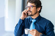 © Joao - Businessman wearing glasses, talking on the phone while holding a cup of coffee and through the window. Happy successful male portrait at the office by the window