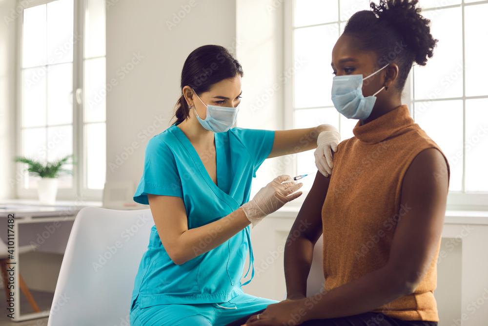 Young African American lady sitting at doctor's office and getting modern Covid 19 shot. Nurse in medical face mask and gloves holding syringe and giving female patient flu vaccine injection in arm - obrazy, fototapety, plakaty 