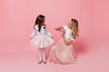 Wall Mural - Dark-haired little girl in cute princess outfit poses for mom taking photo on pink background