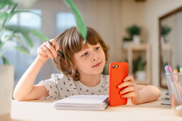 Wall Mural - Back to school. Distance learning online education. Caucasian smile kid boy studying at home with mobile smart phone and doing school homework. Thinking child siting at table with cell phone