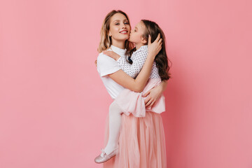 Wall Mural - Lovely mother and daughter in same pink and white outfit posing on isolated background. Woman smiles and holds kid in her arms