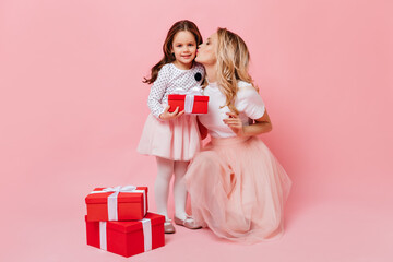 Wall Mural - Loving mother and her daughter pose in same pink and white outfit against isolated background. Kid holds gift while mom kisses her