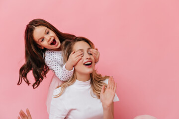 Wall Mural - Emotional little girl closes her eyes to her mother to make surprise. Portrait of girls in white T-shirts on pink background