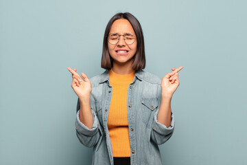 Wall Mural - young hispanic woman smiling and anxiously crossing both fingers, feeling worried and wishing or hoping for good luck