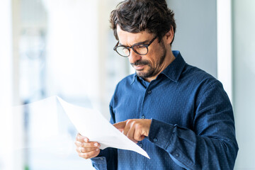 Wall Mural - Business man wearing glasses  reading paper document or contract. Successful male portrait at the office by the window