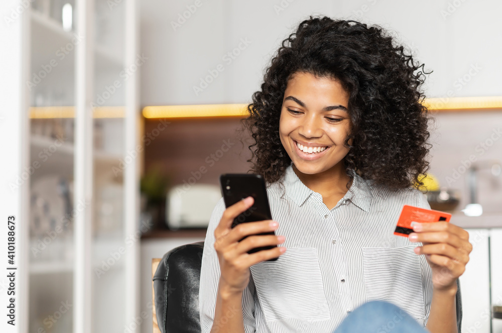 Smiling African American teenage girl with curly hair holding mobile phone, entering credit card number to make an online transaction, mixed-race woman ordering food, doing online shopping from home - obrazy, fototapety, plakaty 