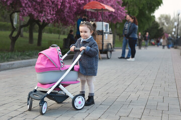 Little girl in a coat walks in the park and rolls a toy stroller with a doll.