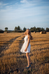 Sticker - Photo of a blond girl wearing a white dress walking in the field of harvested rye. Attractive girl in the agricultural field in the evening