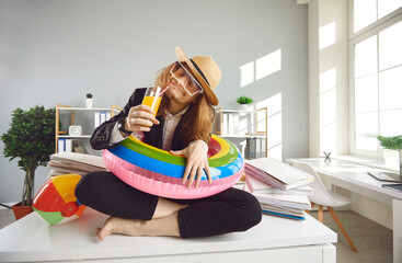 Young office worker in beach sunhat, sunglasses and inflatable swim ring sitting on desk, dreaming eyes closed of summer holiday, thinking of future vacation and sipping drink with happy smile on face