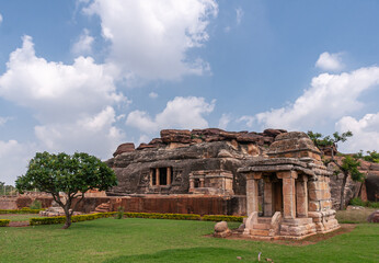 Wall Mural - Aihole, Karnataka, India - November 7, 2013: Ravanaphadi Cave Shiva Temple. Green landscape with brown stone temple buiidings and black rock cave under blue cloudscape.