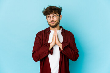 Young arab man on blue background praying, showing devotion, religious person looking for divine inspiration.