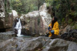 © Sebastian Kanzler/Westend61 - Thoughtful female hiker crouching on rock against waterfall in forest during rainy season
