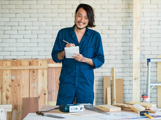 Good looking Asian carpenter wearing coverall dress working for DIY jobs in carpenter room with several kinds of woods and types of equipment. He holding paper notebook for list of things to do