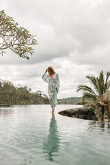 Wall Mural - Happy young woman in long  dress walking on the edge of swimming pool and enjoying summer vacation in daytime