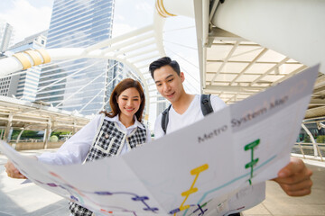 Front view portrait of cute smiling young Asian lover couple travelers holding paper map together on holiday with high building background. Selective focus at the eye with blurred map foreground