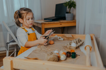 Baby girl decorating easter eggs on white table. Kid decorating eggs with kraft paper, lace and rope. Child cuts the rope with black scissors.