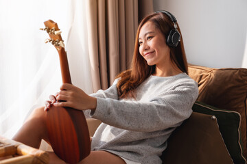 Wall Mural - A beautiful young asian woman holding and looking at an ukulele while enjoy listening to music with headphone at home