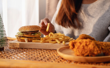 Wall Mural - Closeup image of a woman holding and eating french fries and hamburger with fried chicken on the table at home