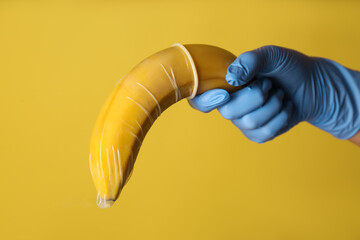 Wall Mural - Doctor holding banana in condom symbolizing male sexual organ on yellow background, closeup. Potency problem