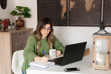 Lifestyle shot of curly blonde hair styled woman sitting on a table while works remote from home using  his laptop and notebook with a mug.