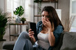© JustLife - Sad woman sitting on the couch, using the phone. Upset woman waiting for a phone call.