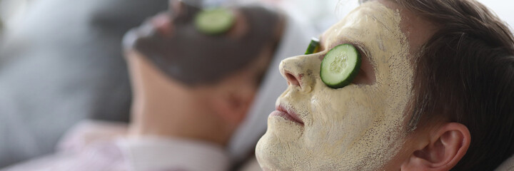 Wall Mural - Close-up of wife and husband wearing cleaning and moisturizing face masks. Relaxed female and male laying on couch. Loving spouse with cucumber slices on eyes. Leisure and cosmetology concept