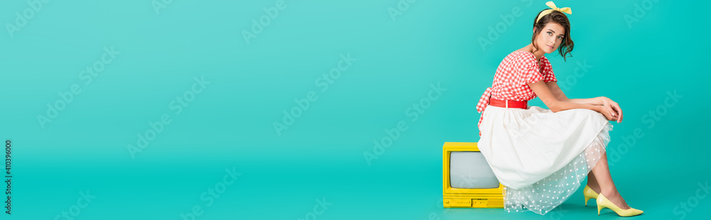 stylish pin up girl looking at camera while sitting on bright yellow retro tv on turquoise, banner - obrazy, fototapety, plakaty 