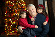 Portrait of the grandfather in a blue sweater with twins granddaughters in red sweaters with long dark hair against the decorated Christmas tree. New Year, family holiday