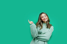 On A Green Background, An Attractive Girl Is Smiling At The Camera, She Points Her Finger At The Background Place For Text. Commercial Photo. Copy Space. Inscription. Advertising