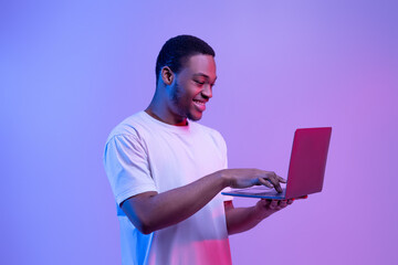 Modern Technologies. Happy Black Man Using Laptop Computer In Neon Light
